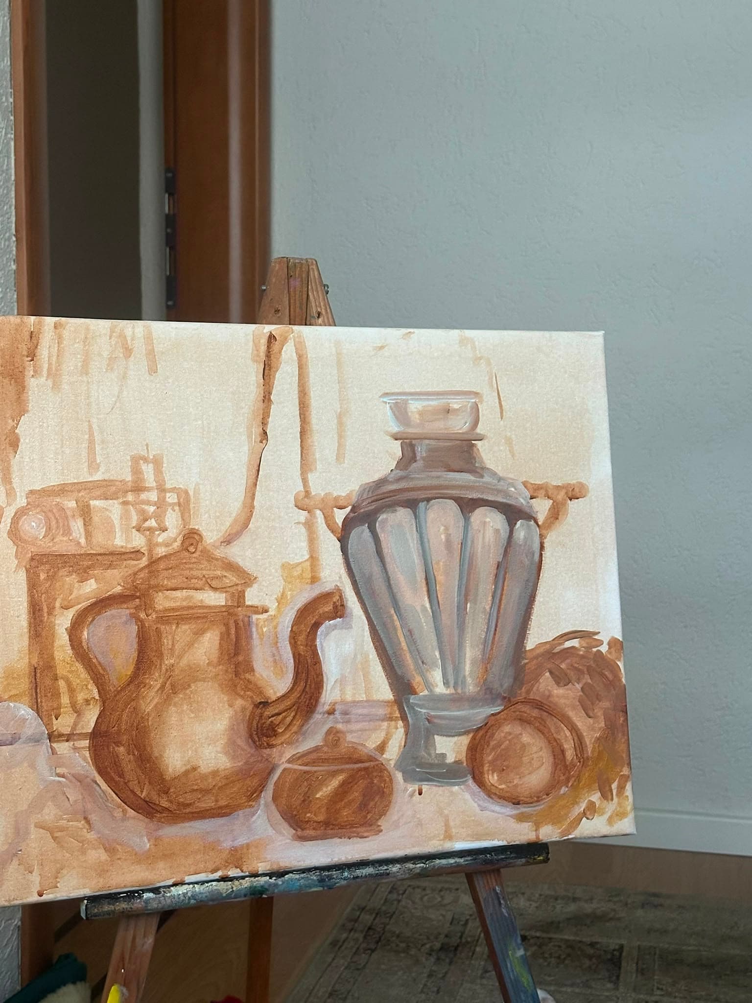 A still life mid block-in on an easel in the studio: a kettle, a small ribbed vase and a bundle of firewood laid in as a rough sepia underpainting, not yet in colour.